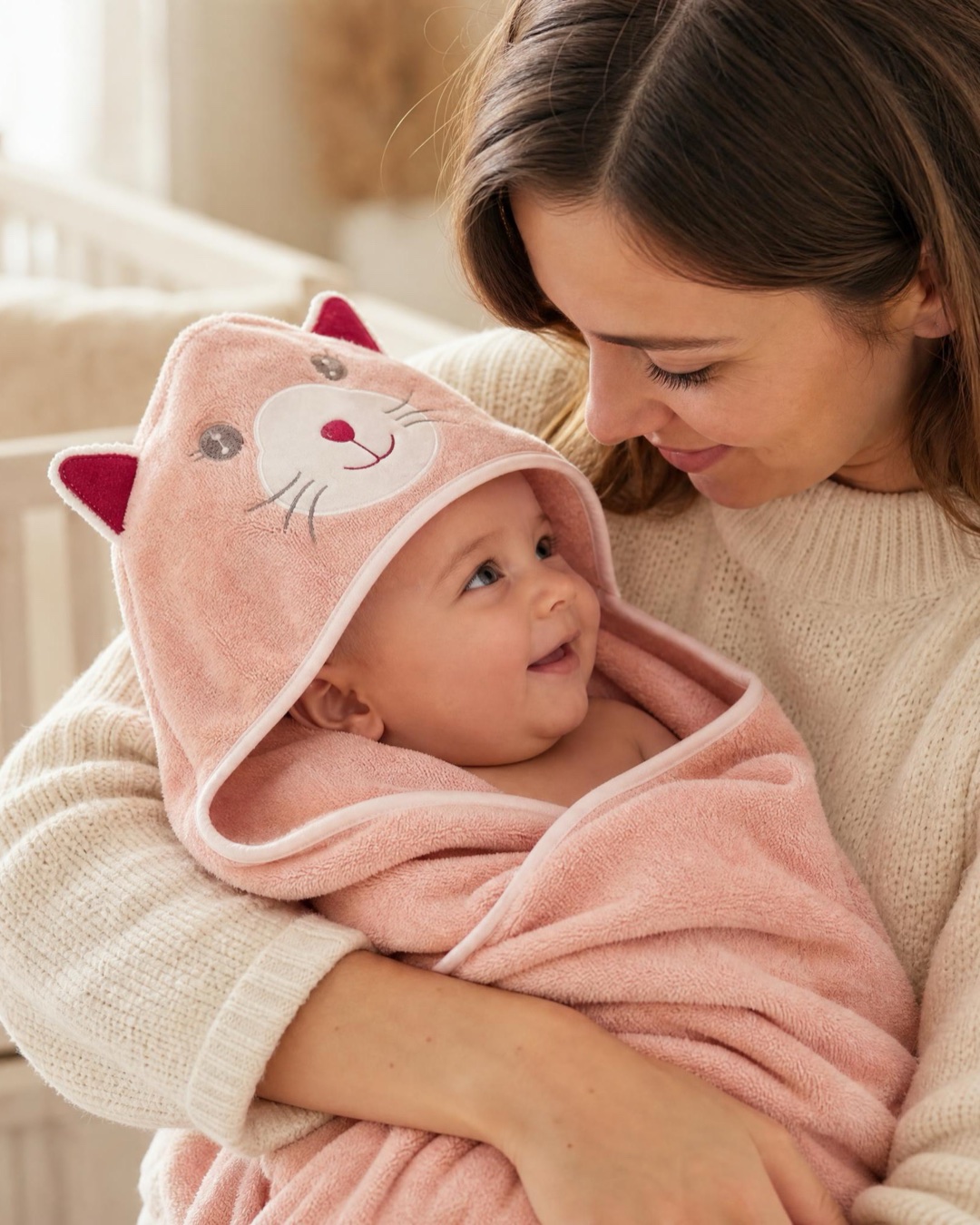 A parent cradling a baby wrapped in a soft pink cat hooded towel beside the crib at bedtime.